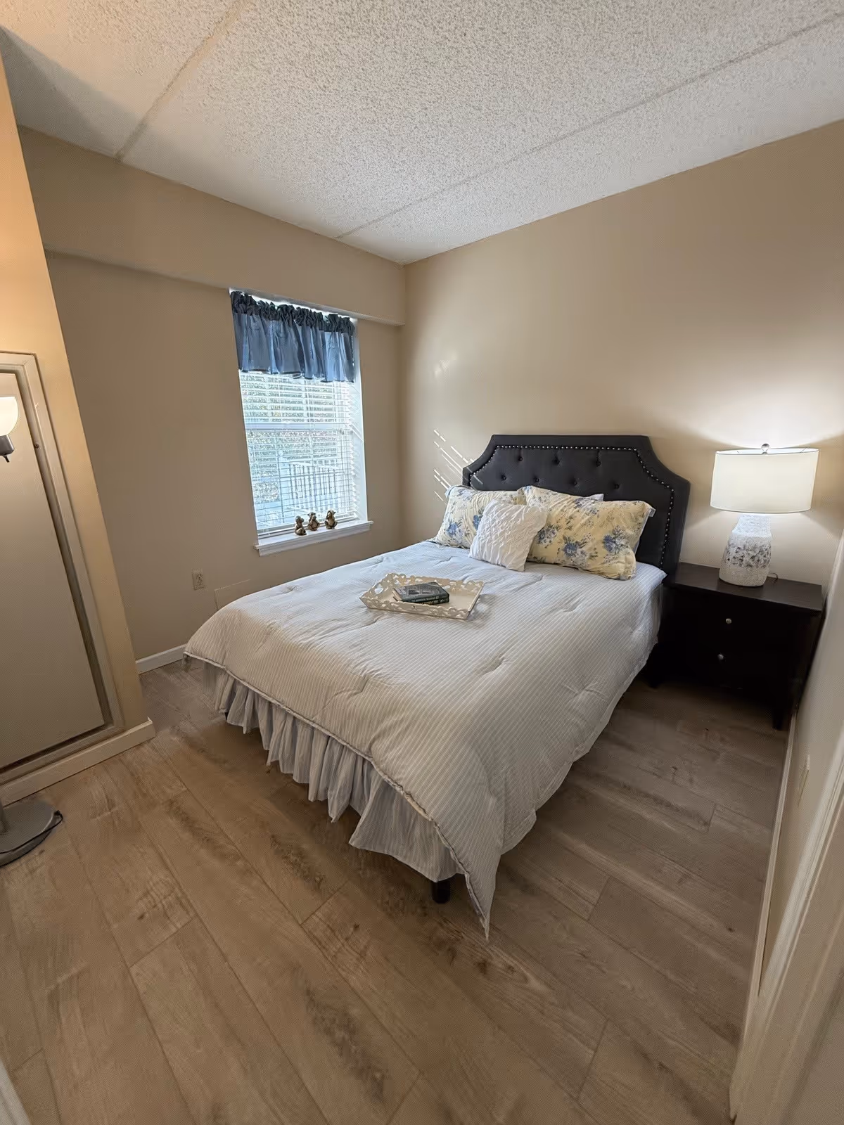 A cozy bedroom with a neatly made bed featuring a tufted dark headboard, white and floral pillows, and a white bedspread. A tray with a book is placed on the bed. There is a window with a blue valance letting in natural light. A dark wooden nightstand with a white lamp is beside the bed. The room has light wood flooring and beige walls.
