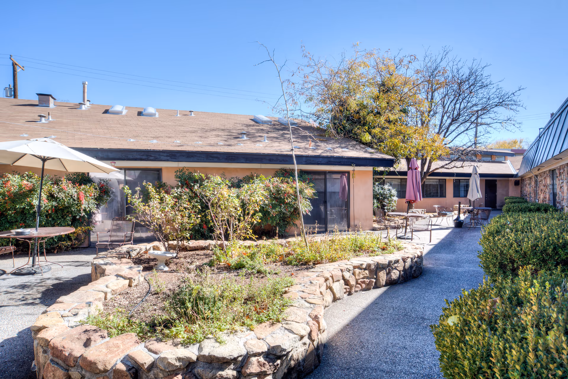 Sunlit courtyard with a raised stone planter, patio tables and umbrellas, shrubs, and a surrounding single-story building.