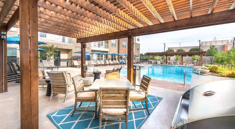 Covered pergola seating and dining area overlooking a swimming pool with lounge chairs and an adjacent apartment building.