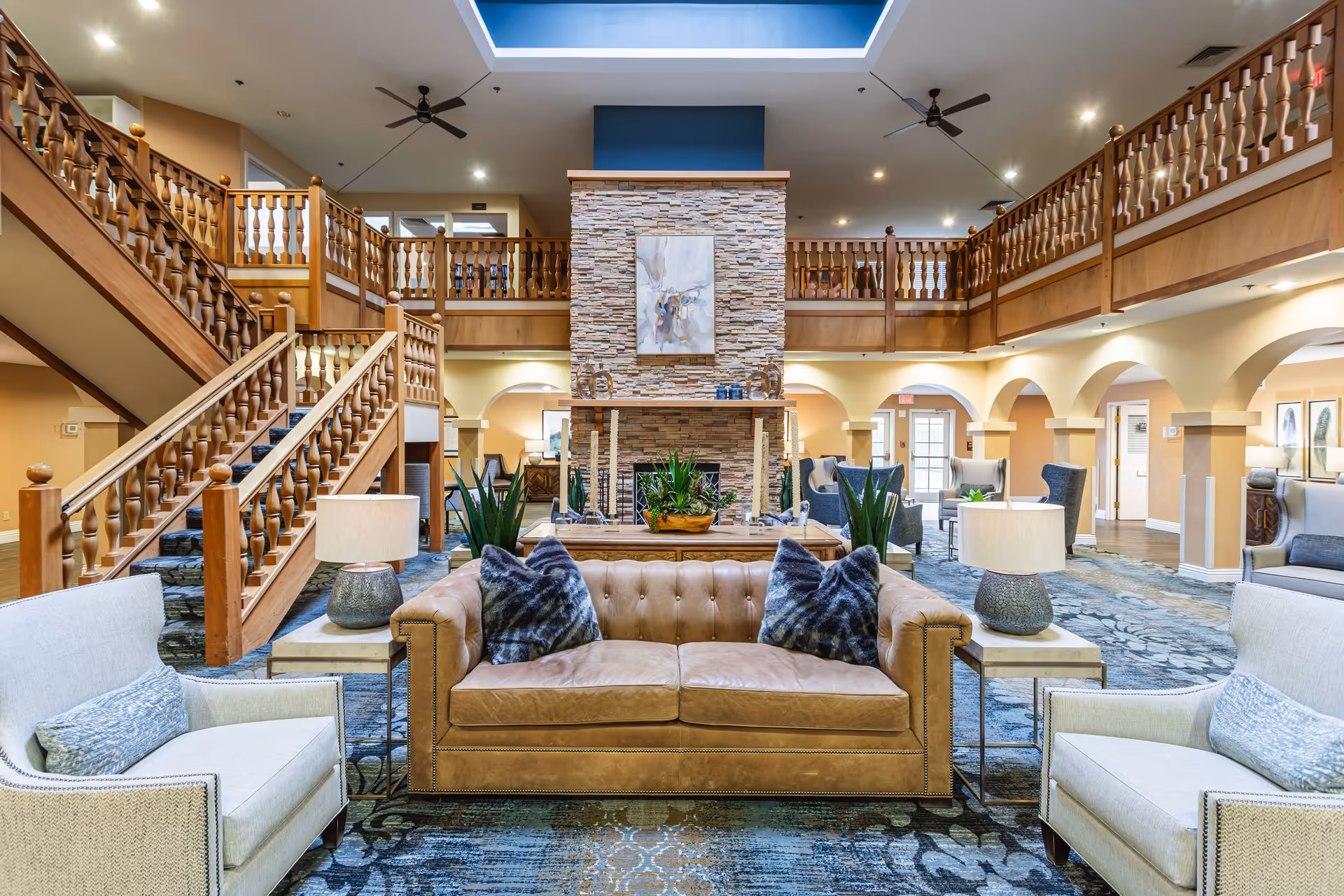 Spacious senior living facility common area with a central stone fireplace, a brown leather sofa with two dark patterned pillows, two beige armchairs with light blue cushions, side tables with lamps, wooden staircase and balcony railings, and a skylight above.