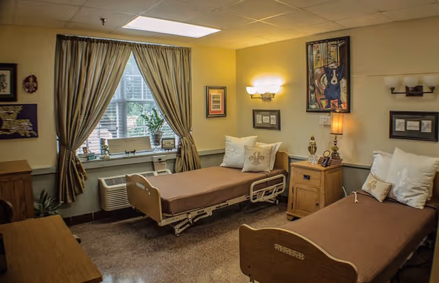 A cozy assisted living bedroom with two hospital-style beds, each with brown bedding and white pillows. The room has beige walls with framed artwork, a window with beige curtains, a wooden nightstand with a lamp and decorative items, and a carpeted floor.