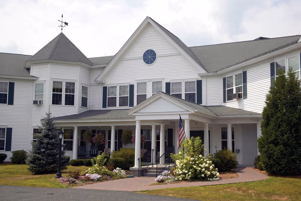 White multi-story senior living building with a columned porch entrance, American flag, and landscaped flower beds.