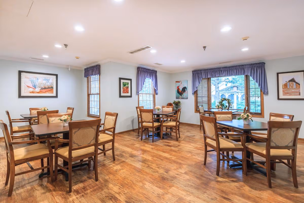 A bright dining room with wooden floors and several tables surrounded by wooden chairs with cushioned seats and backs. The walls are light blue and decorated with framed pictures. Windows with purple valances let in natural light and offer a view of greenery outside.