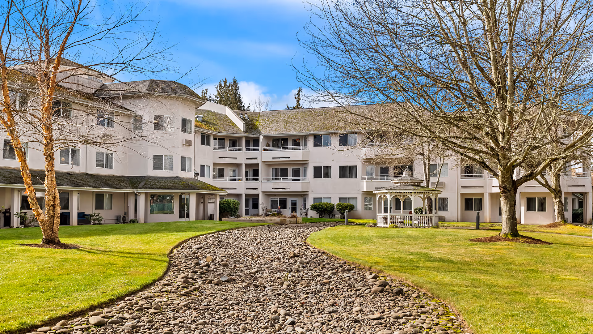 Exterior view of Holiday Sheldon Oaks senior living facility showing a three-story building with multiple windows and balconies. In the foreground, there is a landscaped garden area with green grass, leafless trees, a dry rocky stream bed, and a white gazebo under a blue sky with some clouds.