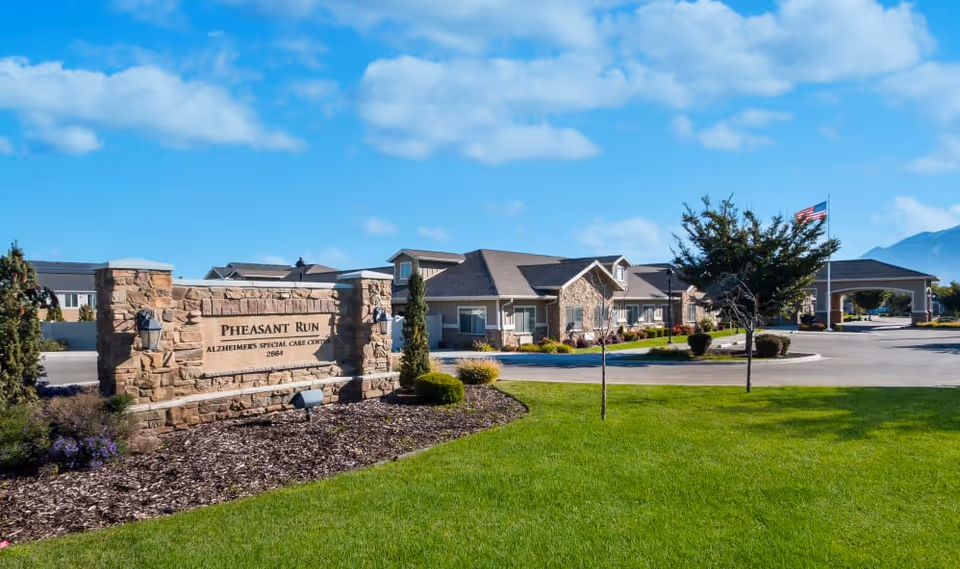 Exterior view of Pheasant Run Alzheimer's Special Care Center with a stone sign in the foreground, well-maintained green lawn, and the building with a covered entrance in the background under a blue sky with scattered clouds.