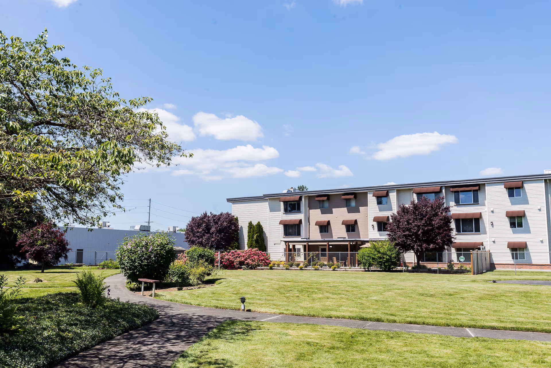 Three-story senior living building with manicured lawn, trees, and a paved walkway under a blue sky.