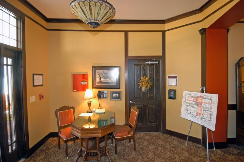 A small seating area inside Webb House Retirement Center with a wooden octagonal table and three upholstered chairs. The walls are painted beige with dark wood trim. A framed picture, a lamp, and a telephone are on the wall above the table. A door with a wreath hangs on the wall behind the table. To the right, there is an easel holding a floor plan. The room has a patterned carpet and a decorative ceiling light fixture.