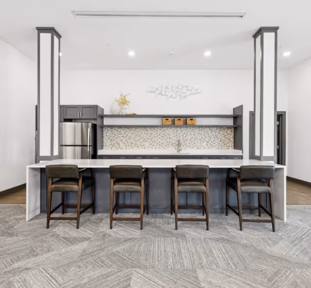 Modern kitchen area with a long white countertop island and four dark cushioned bar stools. The back wall features a stainless steel refrigerator, gray cabinets, a mosaic tile backsplash, and a sink. Decorative items include a vase with dried flowers and three small baskets on a shelf above the sink. The space has a clean, minimalist design with neutral tones and carpeted flooring.