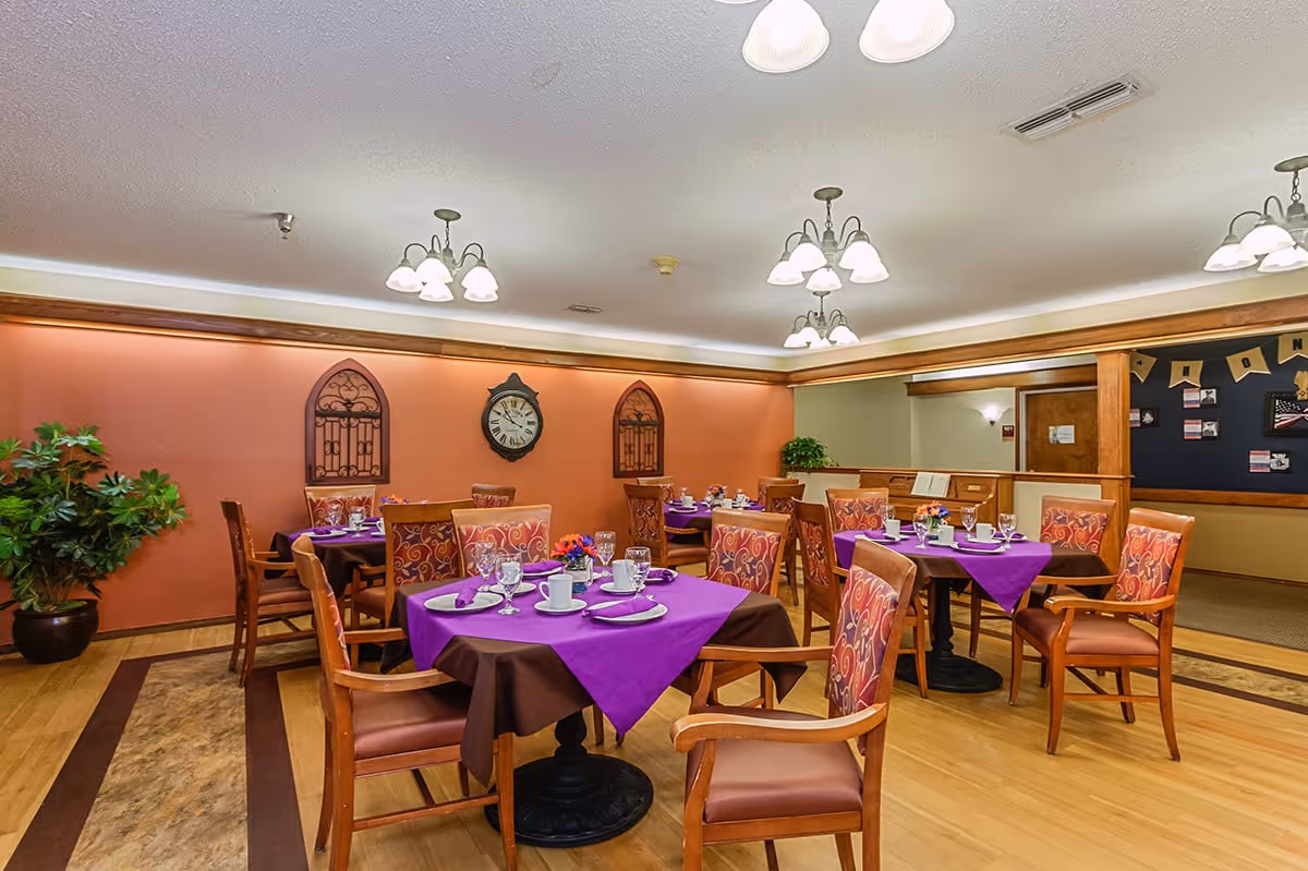 Dining room with several tables set with purple tablecloths, chairs, a wall clock and decorative wall hangings.