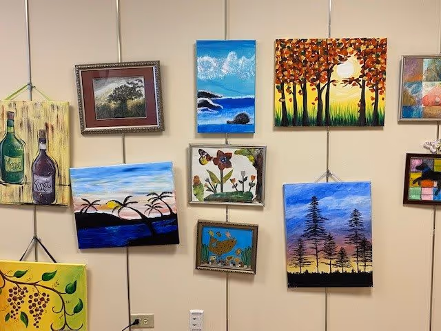 A wall displaying multiple colorful paintings, including landscapes, trees, flowers, bottles, and abstract designs, all hung on a beige wall with visible hanging wires.