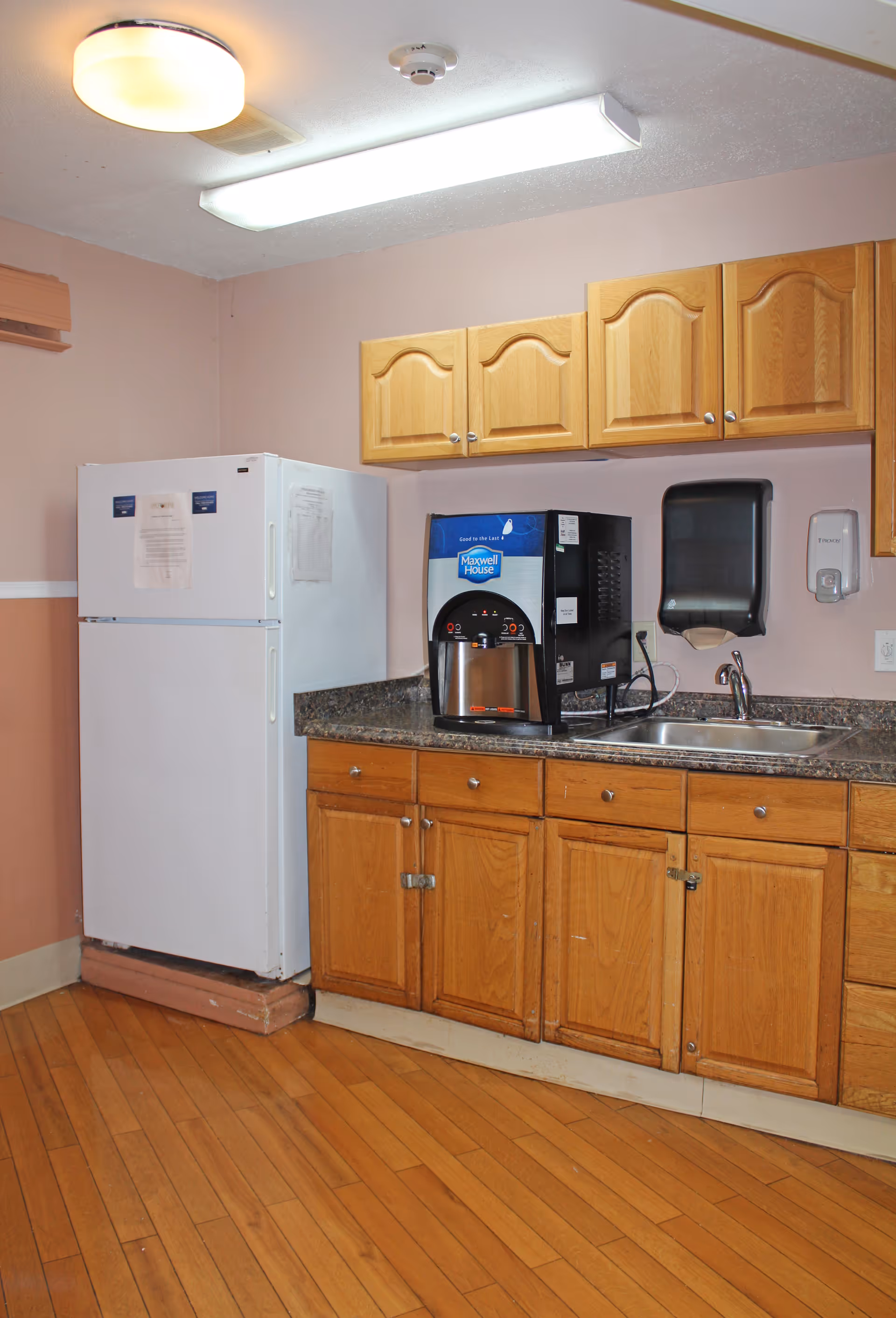 A small kitchen area with wooden cabinets, a granite countertop, a white refrigerator, a coffee machine labeled Maxwell House, a stainless steel sink, a paper towel dispenser, and a soap dispenser mounted on the wall. The floor is wooden and the walls are painted light pink.