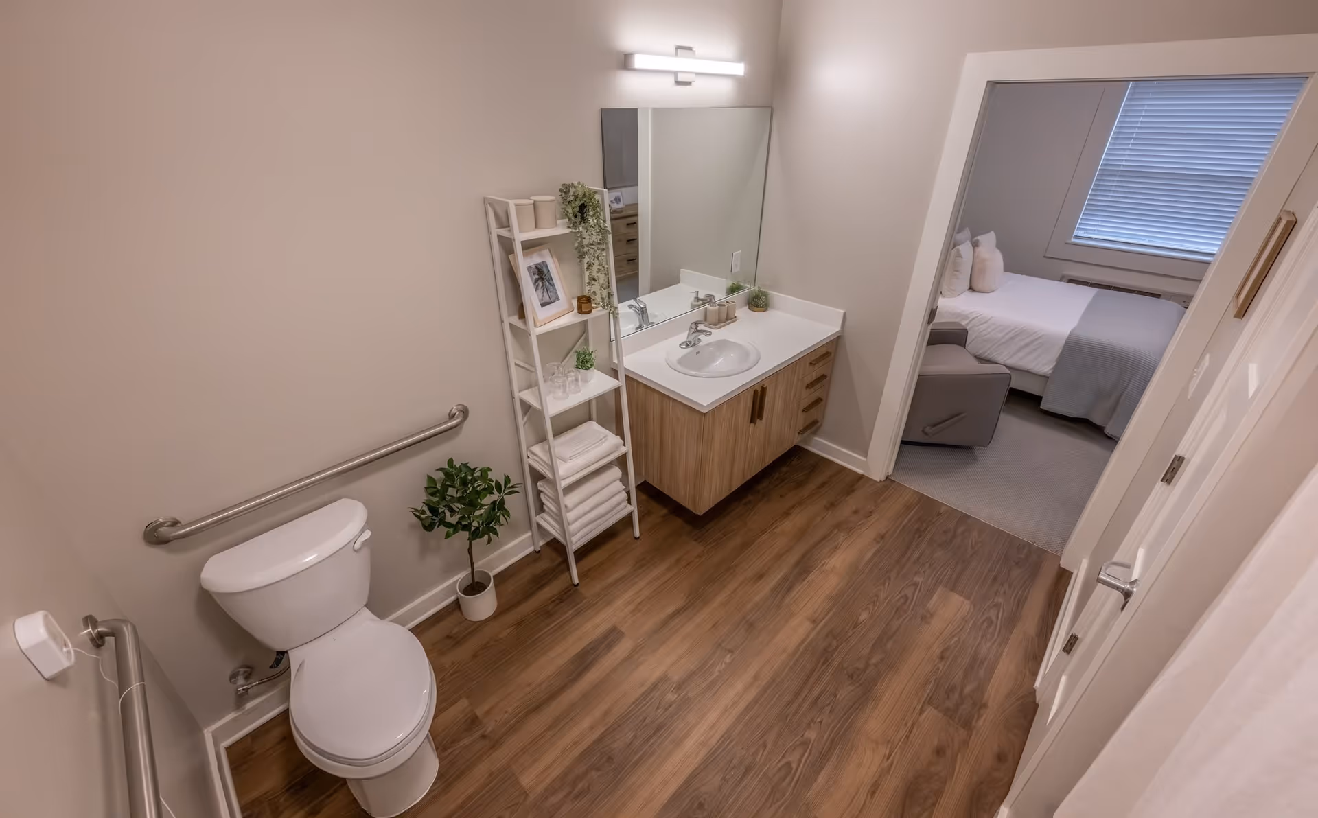 A clean and modern bathroom with a white toilet, a wooden vanity with a sink and large mirror, a white ladder shelf holding towels, plants, and decor, and a small potted plant on the floor. The bathroom has wood-style flooring and beige walls. Through the open door, a bedroom with a bed, pillows, a gray blanket, and a window with blinds is visible.