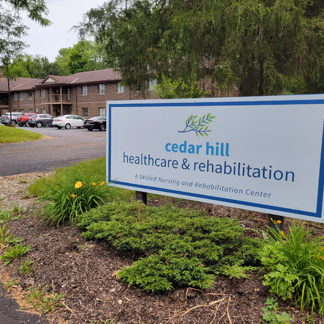Outdoor view of Cedar Hill Healthcare & Rehabilitation facility sign with a landscaped garden in front, showing green shrubs and yellow flowers. In the background, there is a two-story brick building with several parked cars and trees.