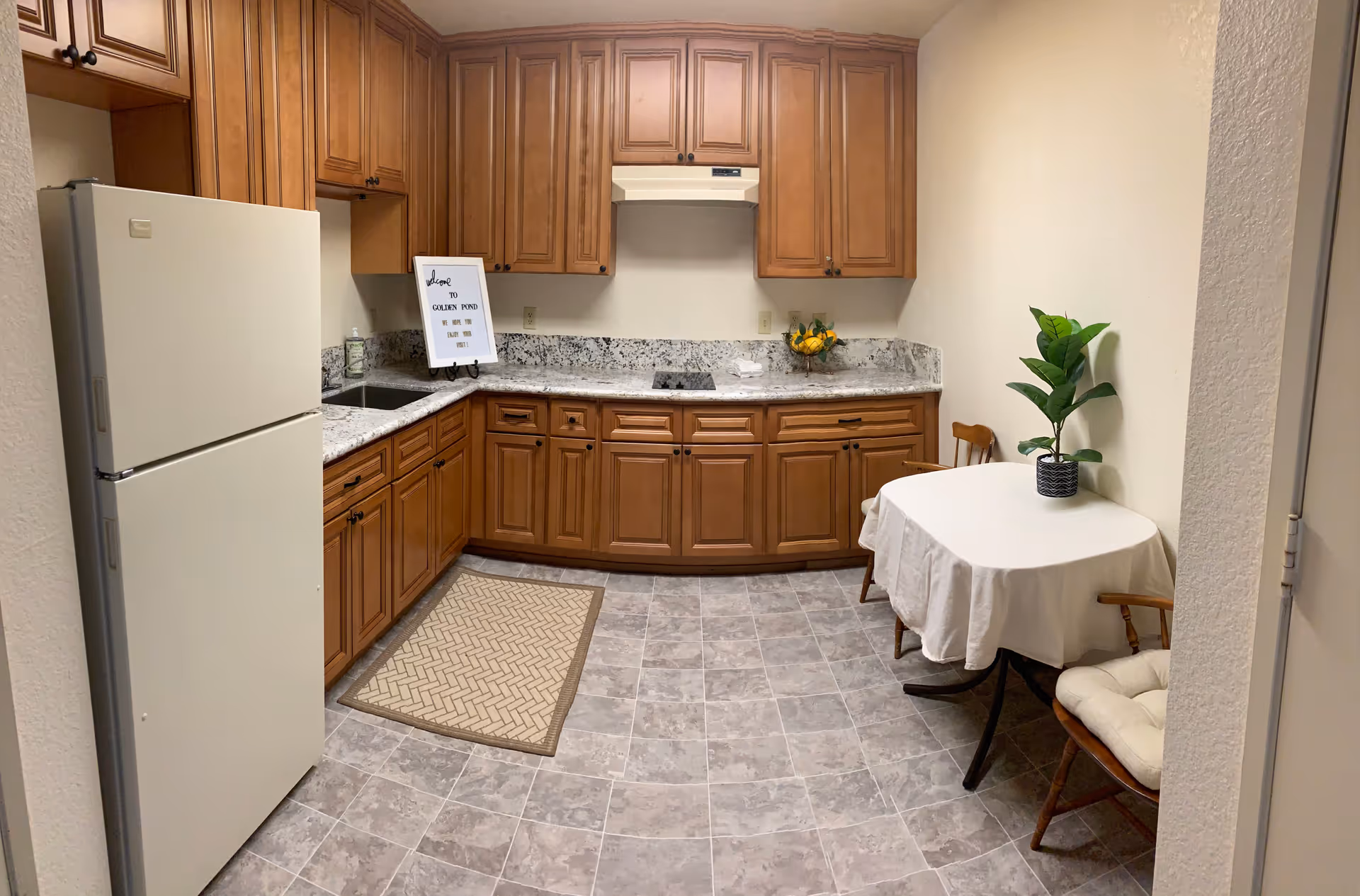 A kitchen with wooden cabinets, a white refrigerator, a granite countertop, and a small dining area with a table covered by a white tablecloth and two wooden chairs. There is a potted plant on the table and a framed sign on the countertop that reads 'Welcome to Golden Pond Retirement Community'. The floor is tiled with a gray pattern.