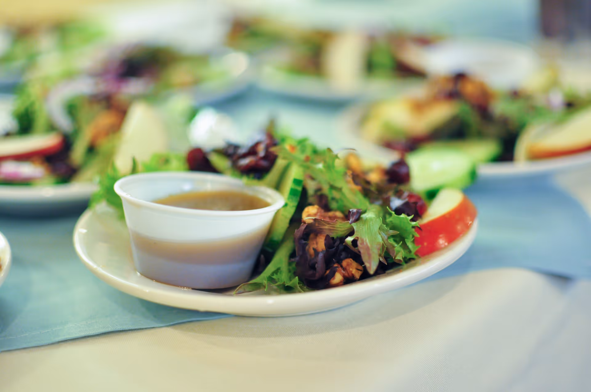 Close-up of a small salad plate with mixed greens, apple slices, walnuts and a cup of dressing on a table.