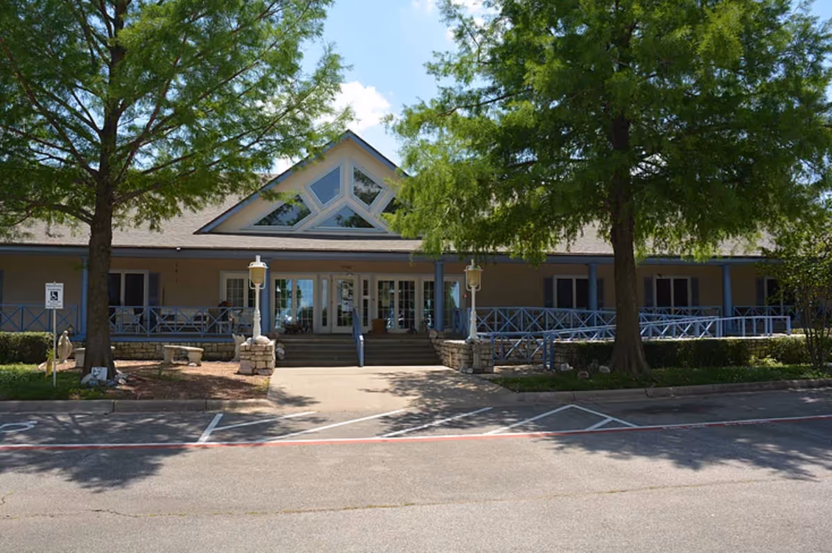 Front entrance of a single-story assisted living building with a covered porch, decorative railing, large triangular windows, and trees in front.