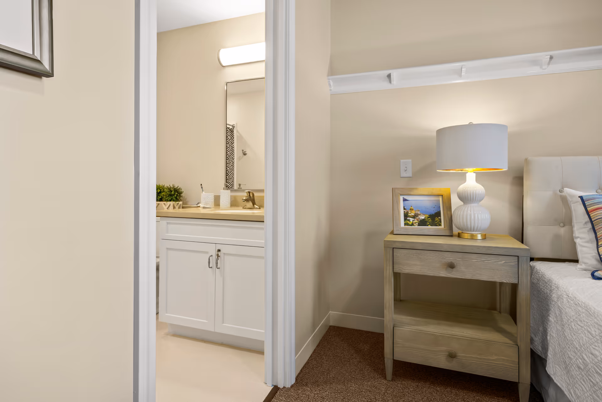 View of a bedroom corner with a wooden nightstand holding a white table lamp and a framed photo, next to a bed with a white quilt and colorful pillows. Adjacent to the bedroom is a bathroom with a white vanity cabinet, a sink, a mirror, and a light fixture above the mirror.