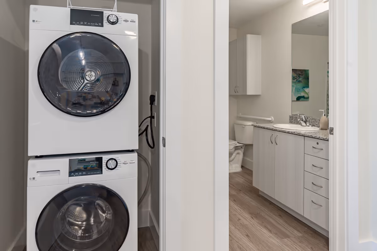 A stacked washer and dryer unit is positioned in a small laundry closet adjacent to a bathroom. The bathroom features a toilet, a vanity with a granite countertop, a sink, a large mirror, and a wall-mounted cabinet. The flooring is wood-style laminate, and the walls are painted light gray.