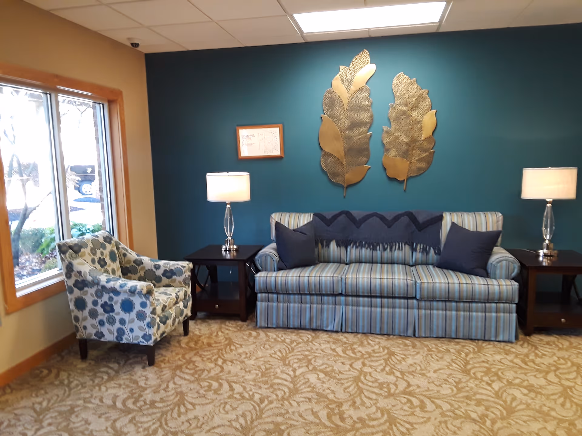 A cozy sitting area with a striped sofa adorned with two dark blue pillows and a matching throw blanket, flanked by two dark wooden side tables each holding a lamp with a white shade. To the left is a floral patterned armchair near a window with a wooden frame. The wall behind the sofa is painted teal and decorated with two large metallic leaf sculptures and a small framed picture.