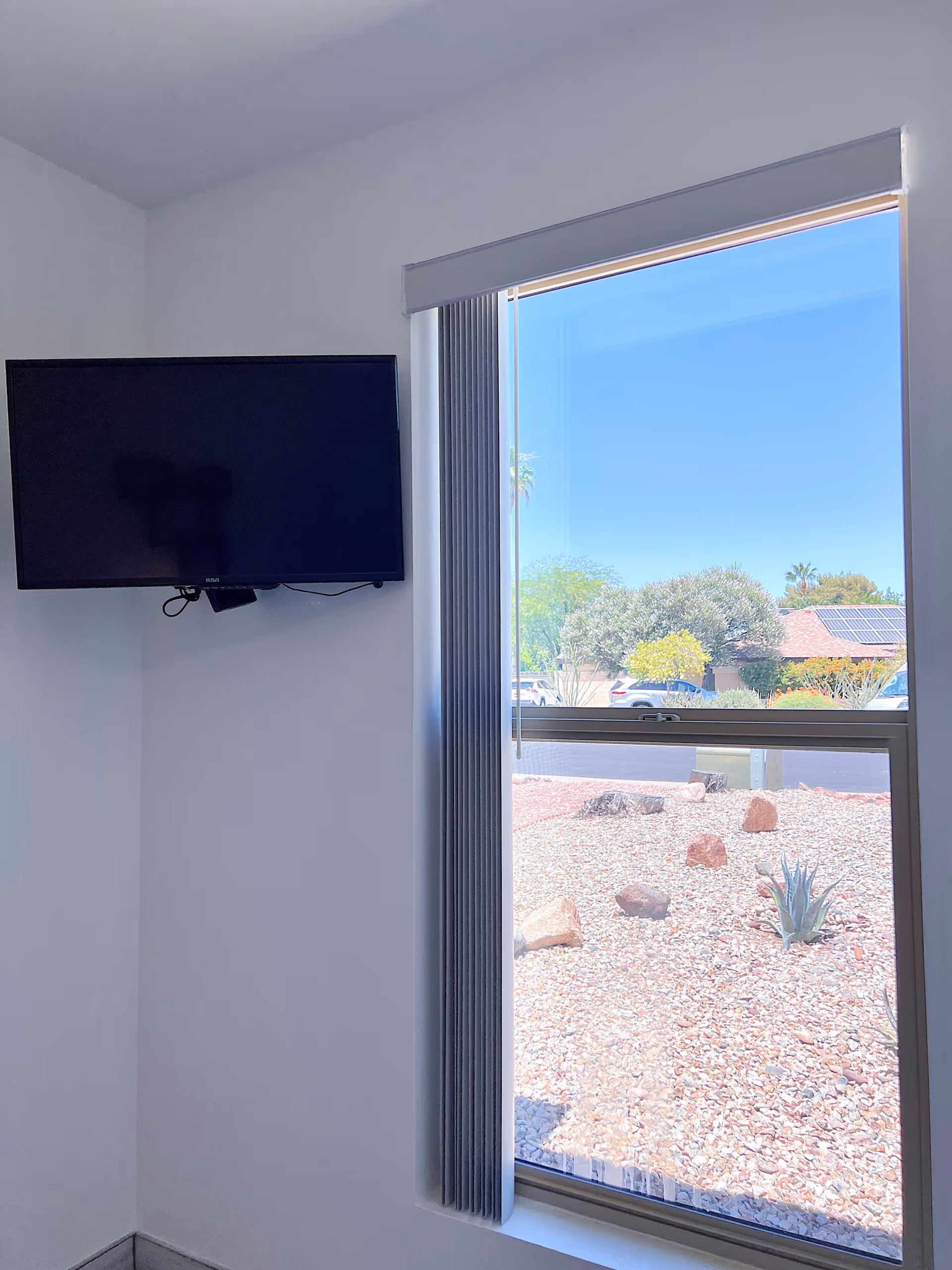 A corner of a room with a wall-mounted flat screen TV on the left and a window with vertical blinds on the right. Outside the window, there is a view of a rocky landscape with some plants and trees, along with a clear blue sky.