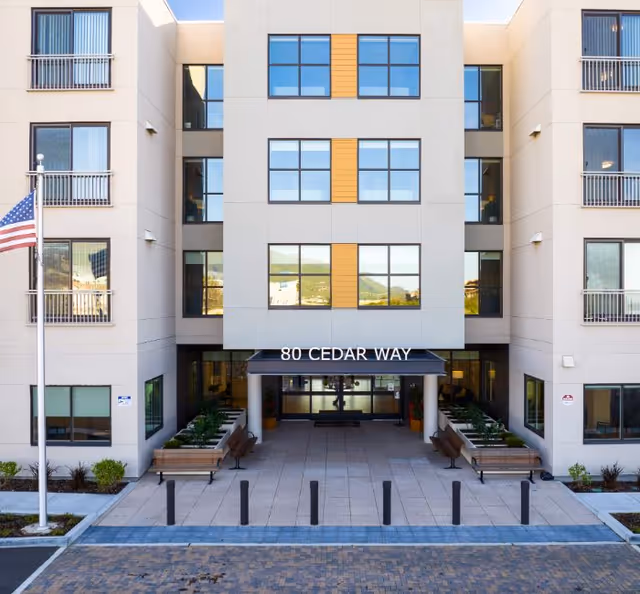 Front exterior view of a multi-story senior living facility building with large windows and a covered entrance labeled '80 Cedar Way'. There are benches and planters along the entrance walkway, an American flag on a flagpole to the left, and a paved driveway in front.
