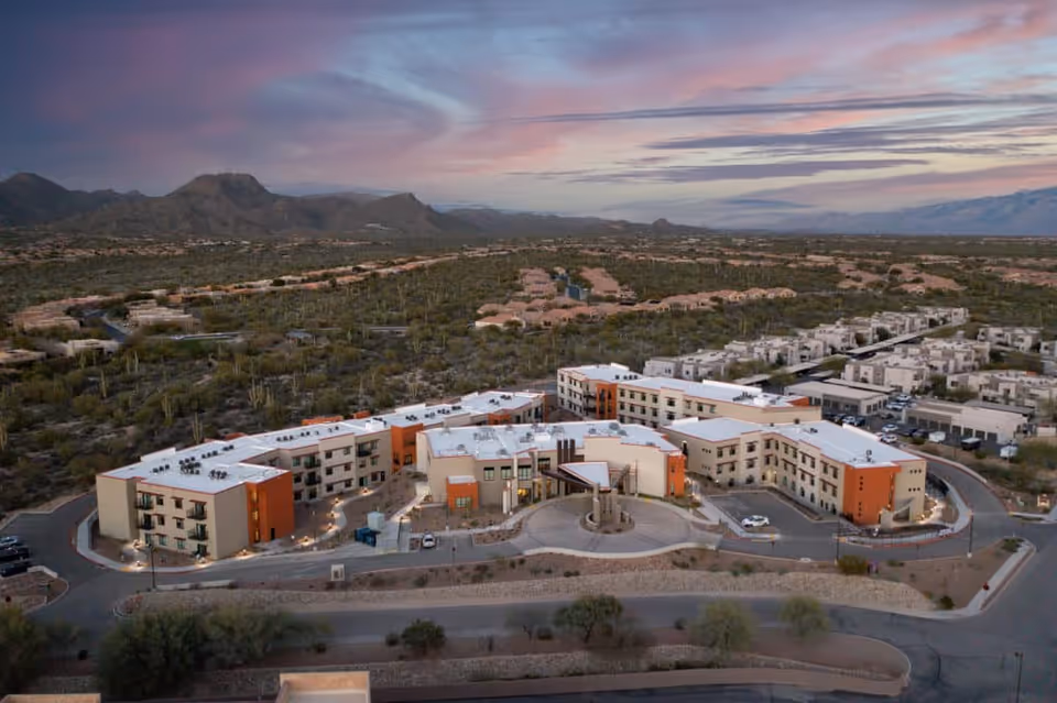Aerial view of La Sonora at Dove Mountain senior living facility surrounded by desert landscape and mountains under a colorful sunset sky.