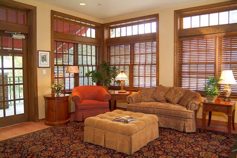 A cozy living room area with a patterned carpet, a beige tufted ottoman in the center, a patterned sofa with cushions, a red armchair, wooden side tables with lamps and plants, and large windows with wooden blinds letting in natural light.