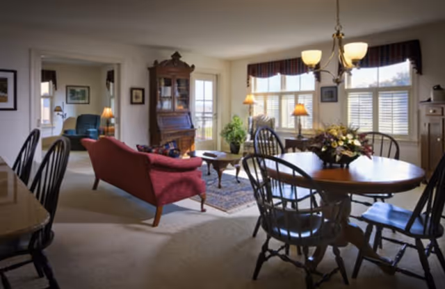 A cozy living and dining area with a round wooden dining table surrounded by chairs, a red upholstered sofa, a wooden coffee table, and a tall wooden cabinet. Large windows with blinds and valances let in natural light, and there are several lamps providing warm lighting. The room has a carpeted floor and an adjacent room with additional seating is visible through an open doorway.
