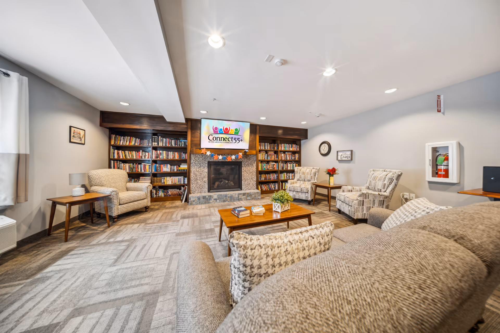 A cozy living room area in a retirement community featuring a stone fireplace with a mounted TV above displaying the Connect55+ logo. The room includes multiple bookshelves filled with books, several upholstered armchairs, a sofa with patterned pillows, a wooden coffee table with books and a plant, and side tables with lamps and flowers. The walls are light gray, and the floor is carpeted with a geometric pattern.