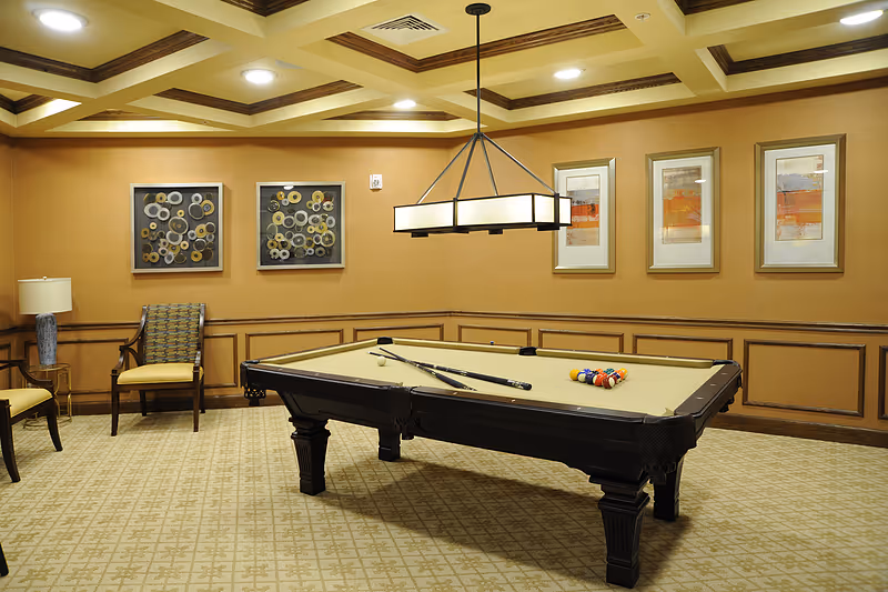 A well-lit room with a beige pool table in the center, two pool cues resting on it, and a rack of billiard balls arranged on one side. The room has patterned carpet flooring, beige walls with wooden paneling, and decorative framed artwork hanging on the walls. There are two chairs and a small table with a lamp in the corner. A rectangular ceiling light fixture hangs above the pool table.