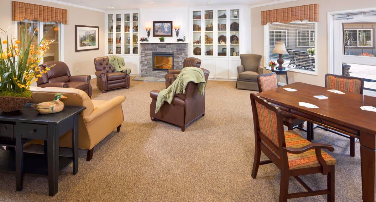 A cozy living room area with several armchairs arranged around a stone fireplace with built-in white cabinets displaying decorative items. There is a wooden table with four chairs on the right side, with playing cards spread on the table. The room has beige carpeting, windows with plaid valances, and a side table with a lamp and flowers.