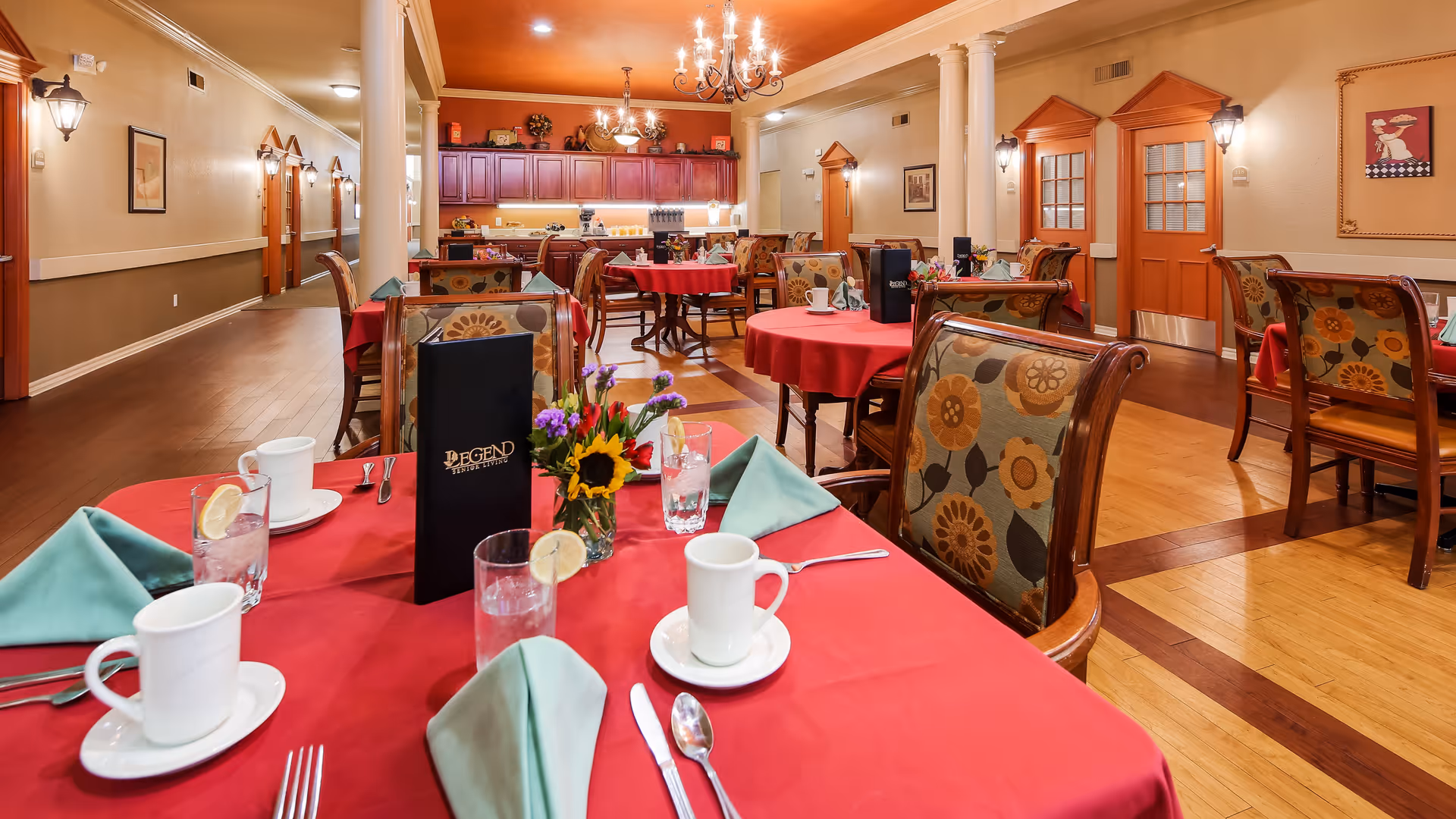 Dining room with multiple round tables covered in red tablecloths, set with white cups, glasses with lemon slices, silverware, and green folded napkins. Chairs have floral patterned upholstery. The room features warm lighting, wooden flooring with a decorative inlay, and a kitchen area with wooden cabinets in the background. A menu with 'Legend At Jefferson's Garden' is visible on the table in the foreground.