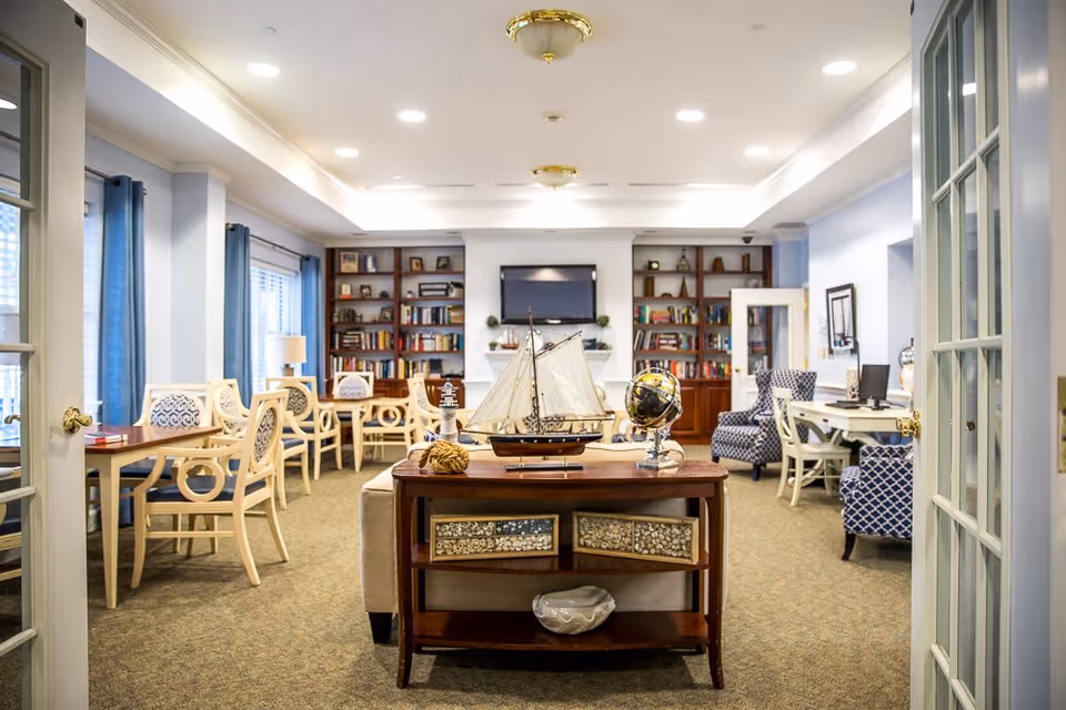 A bright and spacious common area in an assisted living facility featuring a wooden table with a model sailboat and a globe in the foreground. The room has multiple tables and chairs arranged for seating, bookshelves filled with books along the back wall, a flat-screen TV mounted above a fireplace, and large windows with blue curtains letting in natural light. The decor includes patterned armchairs and a computer desk on the right side.
