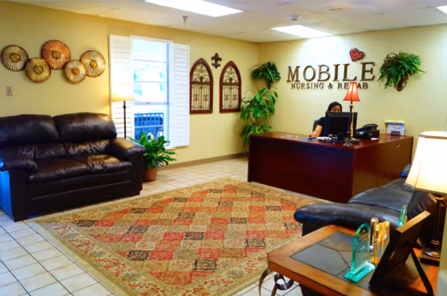 Reception area of Mobile Nursing and Rehabilitation, LLC featuring a dark wooden reception desk with a person working behind a computer, two black leather couches, a patterned area rug, potted plants, wall decorations including circular metal art and arched window-like frames, and a sign on the wall that reads 'MOBILE NURSING & REHAB'.