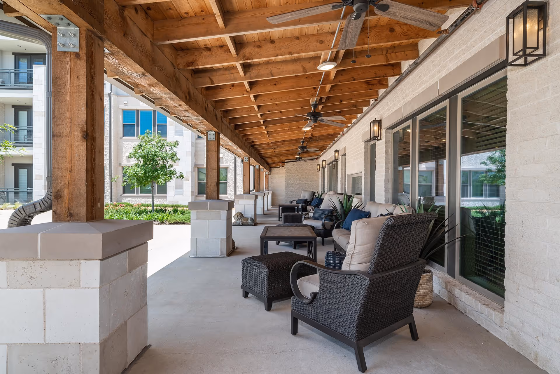 Covered outdoor patio with wicker chairs, sofas and tables under a wooden-beamed roof with ceiling fans alongside the building windows.