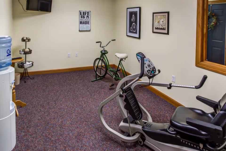 A small exercise room with a recumbent exercise bike and a stationary green bicycle. The room has beige walls with three framed bicycle-themed pictures and a carpeted floor. There is a water cooler and a tiered stand with towels in the corner, and a wall-mounted TV in the upper left corner.