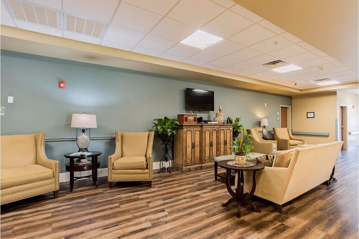 A cozy living room area in a senior living facility with beige upholstered armchairs and a sofa arranged around wooden side tables. A wooden cabinet with decorative plants and a flat-screen TV mounted above it is against a green accent wall. The room has wood-patterned flooring and a drop ceiling with recessed lighting.