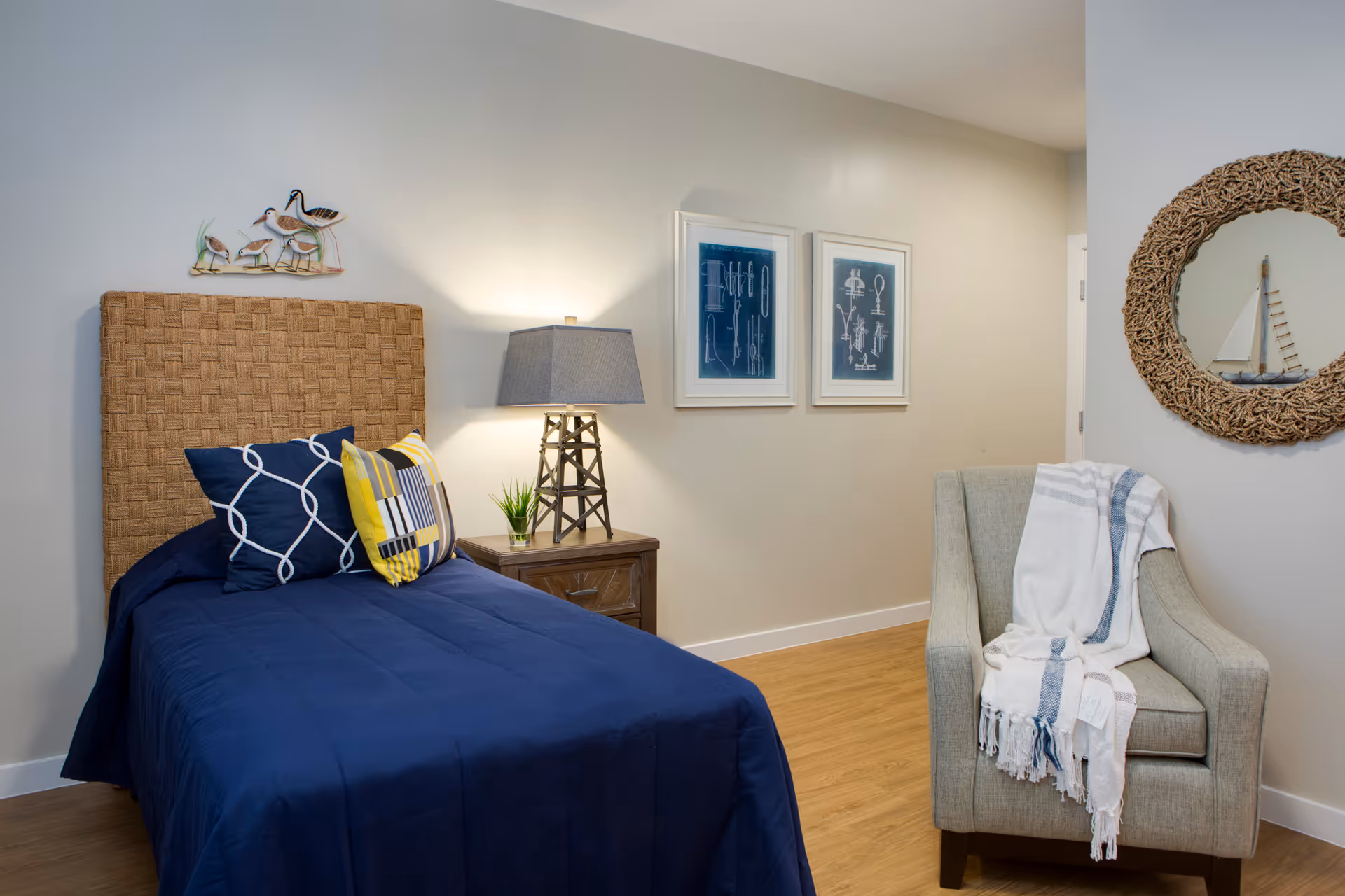 A cozy bedroom with a single bed featuring a woven headboard and navy blue bedding with decorative pillows. Next to the bed is a wooden nightstand with a lamp and a small plant. On the wall above the bed is a metal wall art of birds. Two framed blueprints hang on the adjacent wall. A comfortable armchair with a white and blue striped throw blanket is positioned near a round mirror with a woven frame.