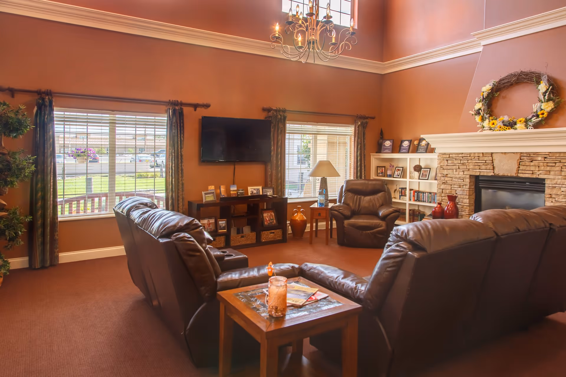 A cozy living room with brown leather sofas and an armchair arranged around a stone fireplace decorated with a floral wreath. The room has large windows with curtains, a wall-mounted TV, a wooden side table with a lamp, and a bookshelf with framed photos and books. A chandelier hangs from the ceiling, and there is a small table with a drink and magazines in the foreground.