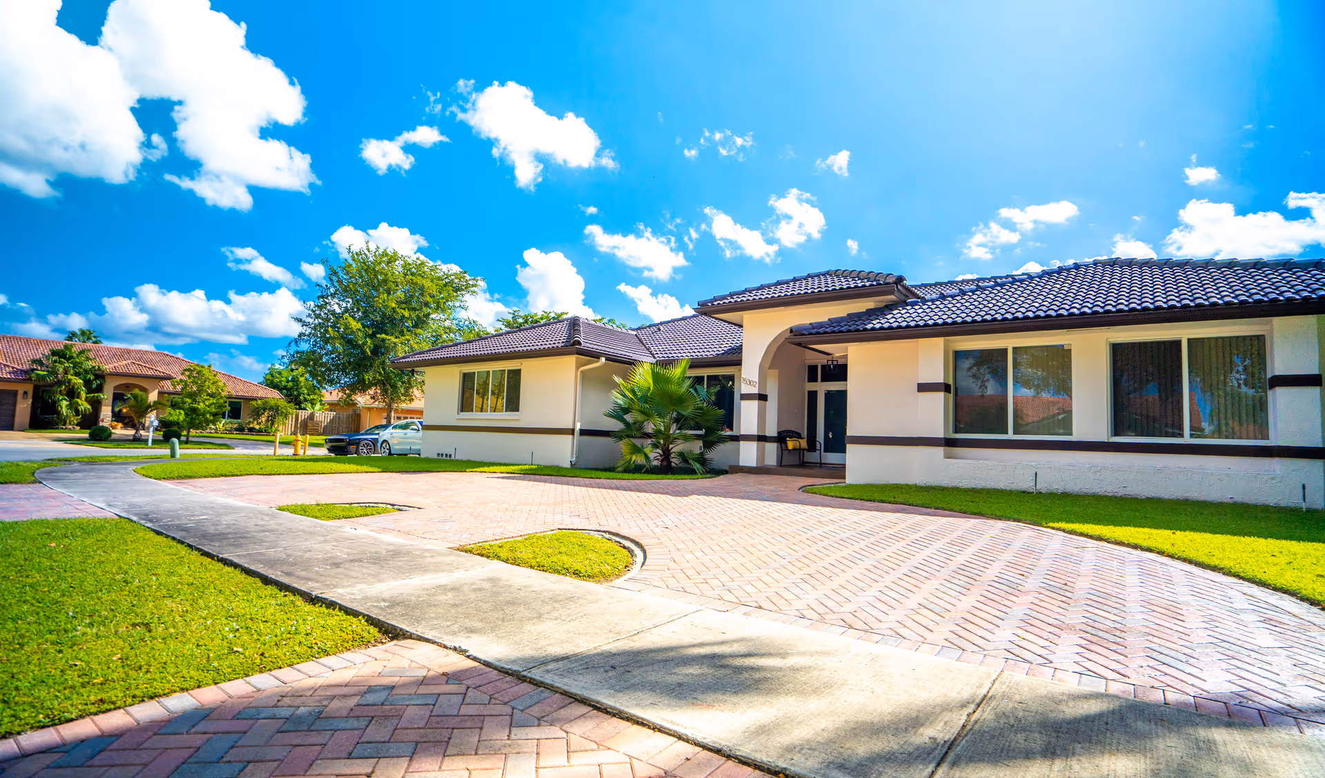 Exterior view of a single-story residential building with a tiled roof, large windows, and a paved driveway under a bright blue sky with scattered clouds. The surrounding area features green lawns, trees, and a sidewalk.