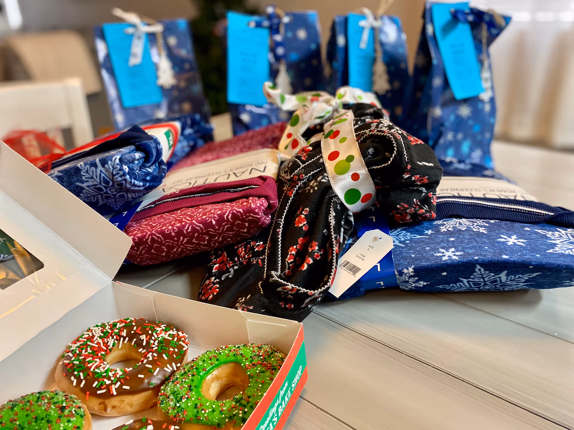 A close-up view of a table with holiday-themed donuts in an open box in the foreground and several folded pairs of festive socks tied with ribbons in the background.