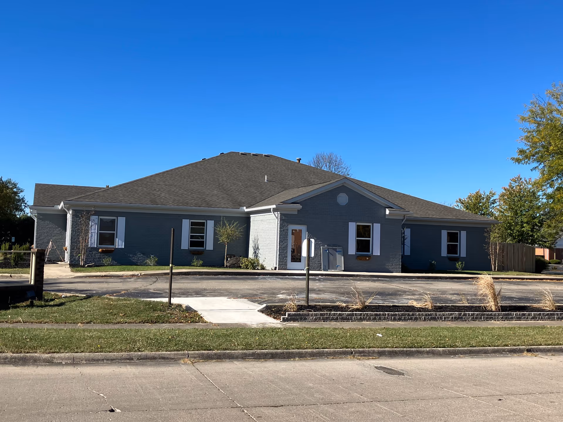 Exterior view of a single-story gray brick building with white window shutters and a dark shingled roof under a clear blue sky. The building is surrounded by a small landscaped area with grass, small trees, and shrubs, and there is a paved driveway and sidewalk in front.