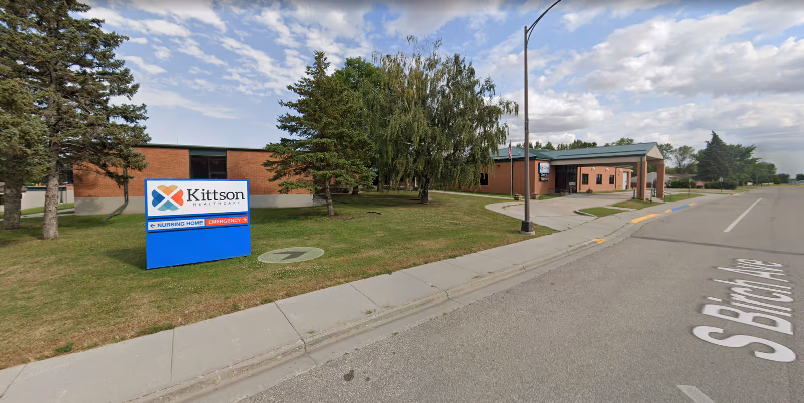 Front exterior of Kittson Healthcare nursing home with sign, lawn, trees, and a covered entrance on S Birch Ave.
