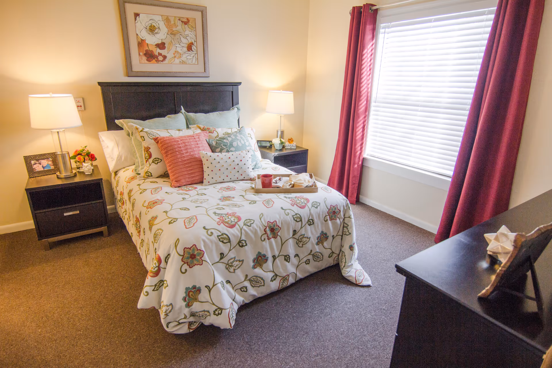 Bright furnished bedroom with a bed dressed in floral bedding, two nightstands with lamps, and a window with red curtains.