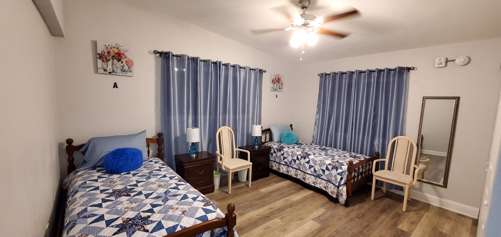 A bedroom with two single beds, each with a blue and white patterned quilt and a round decorative pillow. Between the beds are two dark wooden nightstands with lamps. Two chairs are placed near the beds. The room has light-colored walls, two windows with blue curtains, a ceiling fan with lights, a wall mirror, and wooden flooring.