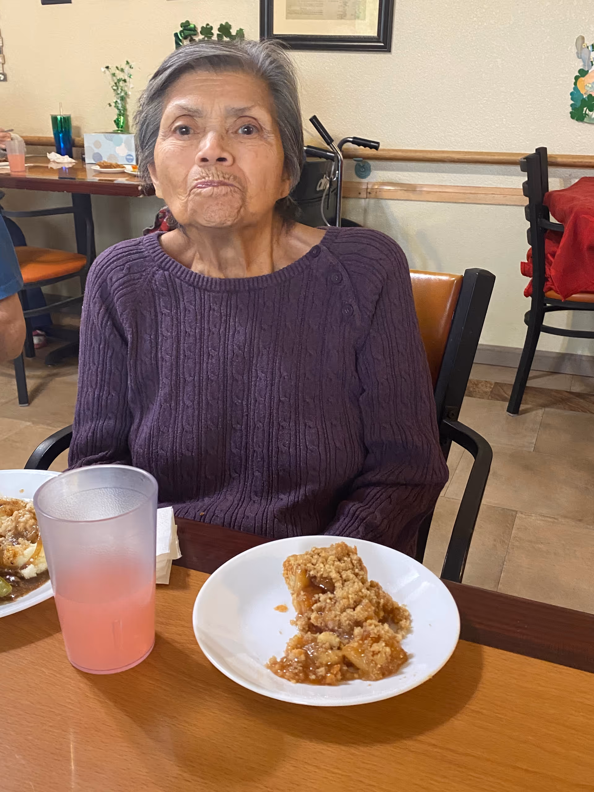 An elderly woman wearing a purple sweater is sitting at a dining table with a plate of dessert and a glass of pink beverage in front of her. The background shows chairs, a walker, and some wall decorations in a dining area.