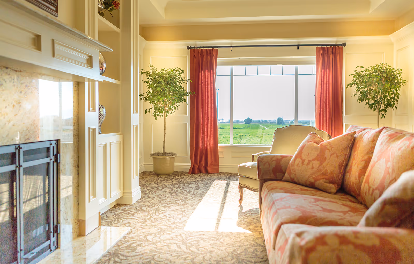 A bright and cozy living room with a patterned sofa and armchair, two potted plants, a large window with red curtains overlooking a green field, and a fireplace with a marble surround and built-in shelves.