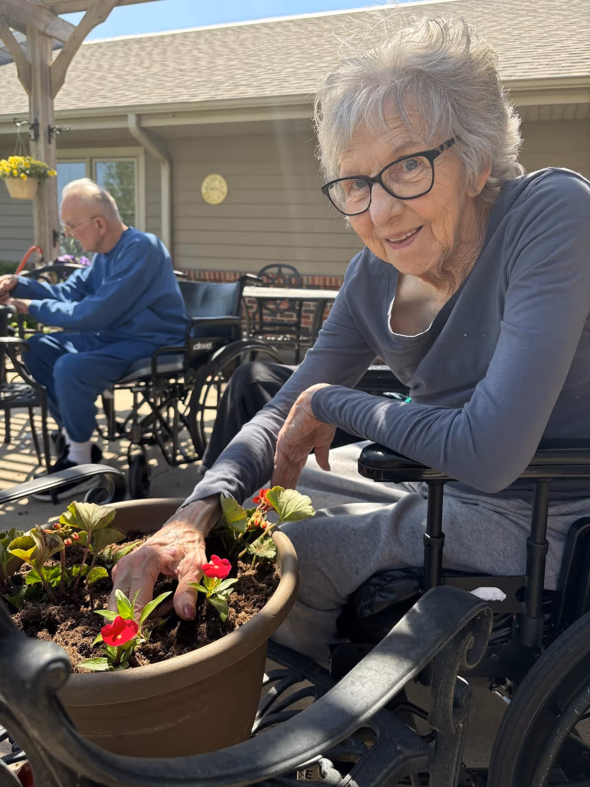 An elderly woman in a wheelchair is smiling and planting red flowers in a large pot outdoors. In the background, an elderly man in a wheelchair is also engaged in an activity under a wooden pergola, with a building wall and windows visible behind them.