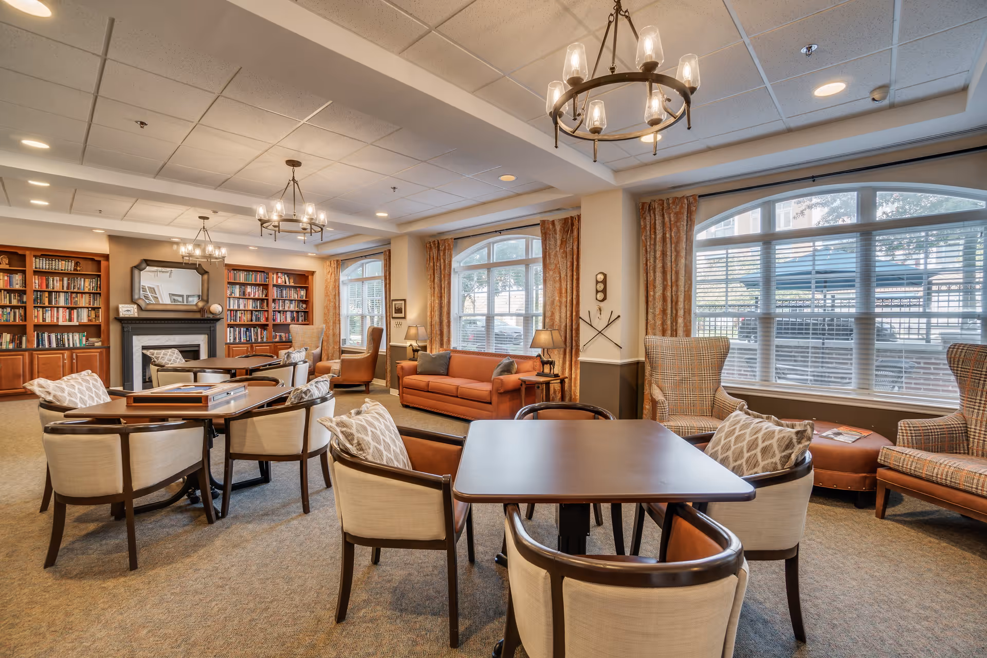 A spacious and well-lit living room area in an assisted living facility featuring multiple seating arrangements including cushioned chairs around tables, armchairs, and a sofa. The room has large arched windows with curtains allowing natural light, bookshelves filled with books, a fireplace with a mirror above it, and elegant chandeliers hanging from the ceiling.