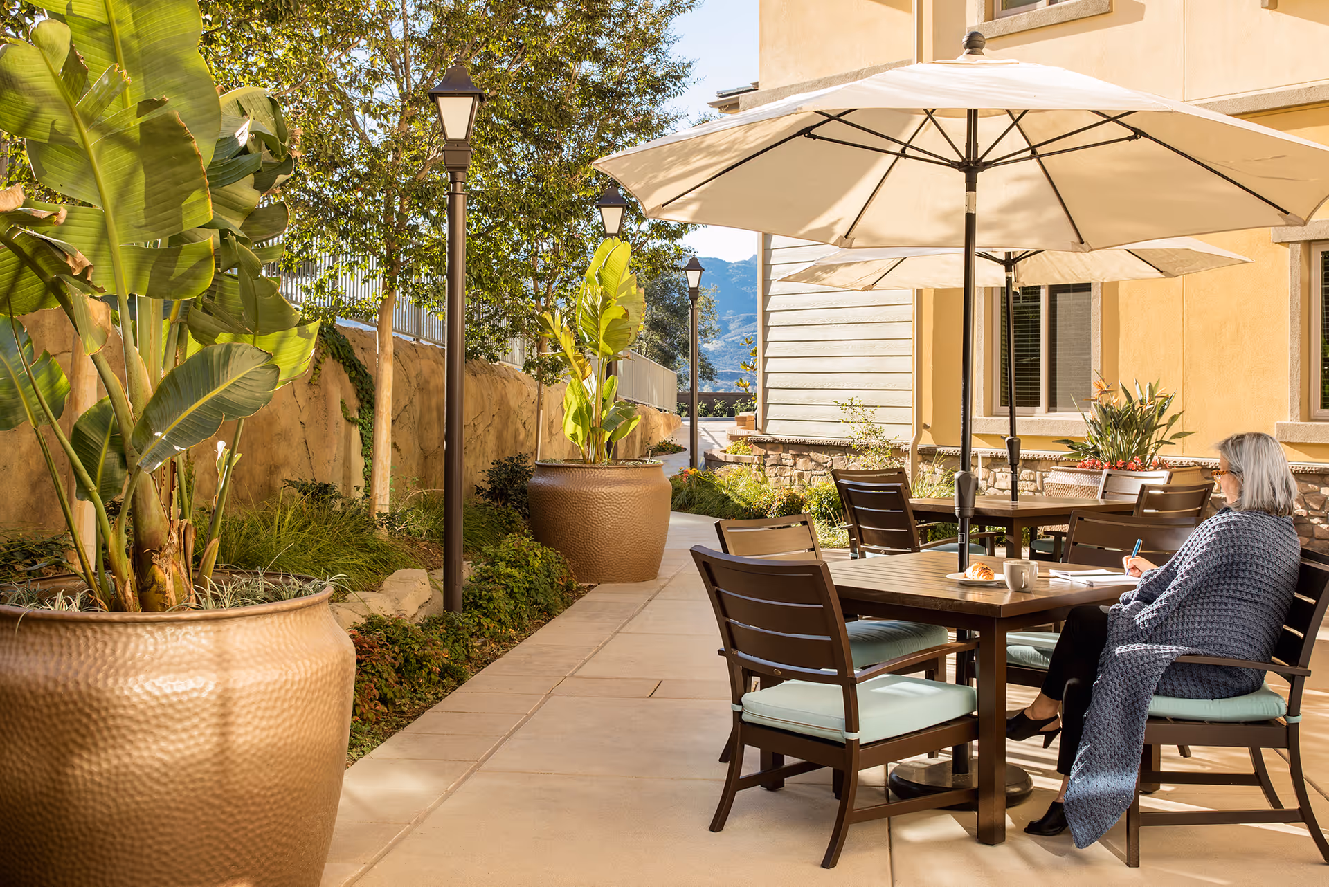 Outdoor patio area at Sage Mountain with large potted plants, several tables with chairs, and umbrellas providing shade. A woman with gray hair is sitting at one of the tables, wrapped in a gray shawl, writing on a notepad with a cup and a pastry on the table.
